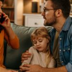 Sad little girl sitting between parents on sofa while her mother talking on phone_s