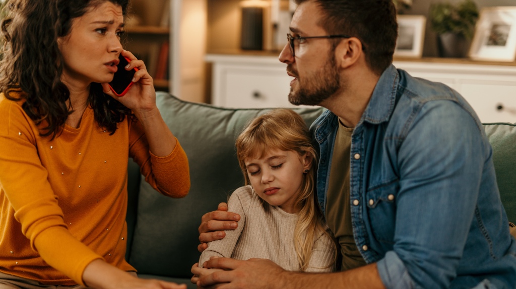 Sad little girl sitting between parents on sofa while her mother talking on phone
