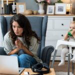 A concerned woman clasps her hands while looking at a laptop, seated near a baby in a high chair_s