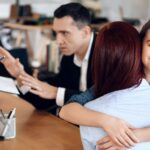 A mother embraces her daughter, who smiles at the camera, while a man gestures toward a lawyer_s