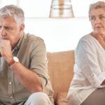 An older couple sits apart on a sofa, appearing tense and emotionally distant_s